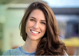 A young woman with long brown hair smiles at the camera outdoors, wearing a light blue top.