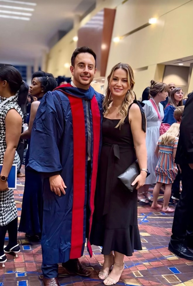 A man in a graduation gown and a woman in a black dress stand together and smile, surrounded by people in a busy indoor setting.